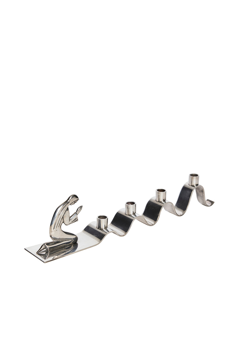 Adventsljusstake av Walter Andersson för Ystad Metall i krom.  I ena änden återges en stiliserad knäböjande figur i bön med subtila art déco-influenser.