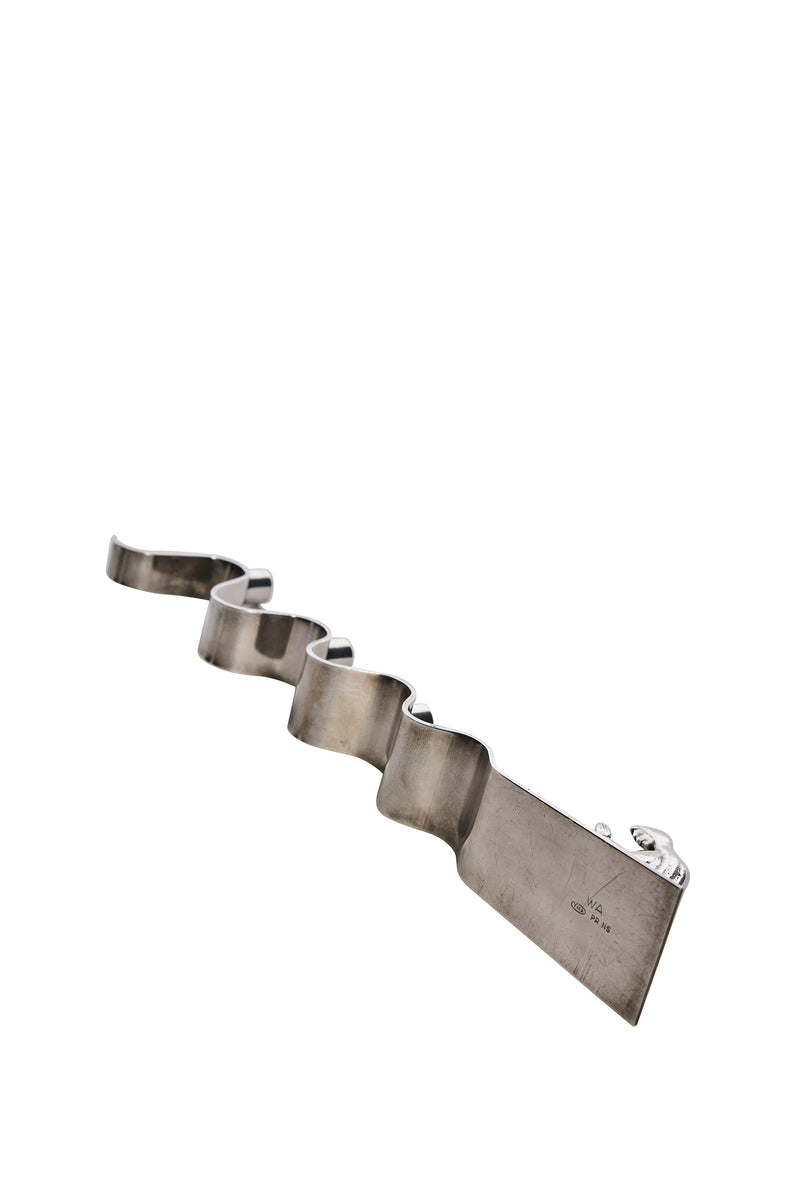 Adventsljusstake av Walter Andersson för Ystad Metall i krom.  I ena änden återges en stiliserad knäböjande figur i bön med subtila art déco-influenser.